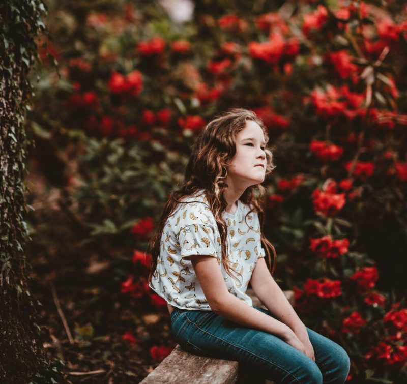 little girl sitting on a bench with red flowers behind her