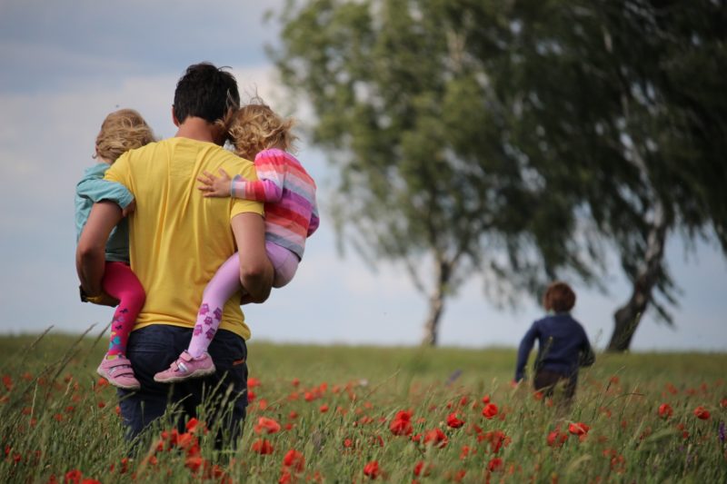 A parent walks through a poppy field holding two children. A third child is running ahead of them.