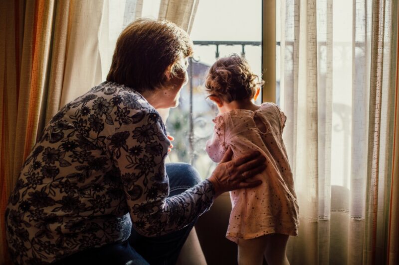 An older woman places a hand on a toddler as they look out the window together
