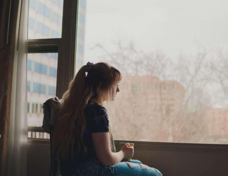 A woman sitting looking out of a window holding a mug in her hand.